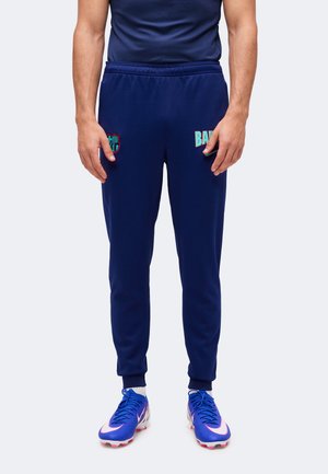 Homme portant un pantalon de sport bleu foncé de Barcelone et des crampons de football bleus assortis, debout sur un fond clair uni.