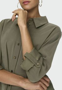 Abito camicia verde oliva con colletto, bottoni sulle spalle e maniche arrotolate. Tessuto morbido e design strutturato, con dettagli metallici.