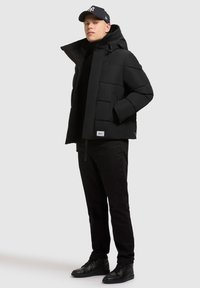 Chaqueta de plumas negra con un cuello alto, patrón acolchado y capucha ajustable. Combinada con pantalones negros y zapatillas, luciendo una gorra de béisbol.
