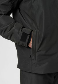 Schwarze wasserdichte Jacke mit weicher Textur, ausgestattet mit einem verstellbaren Klettverschluss-Manschette und einer seitlichen Handtasche. Leicht taillierte Passform.
