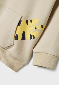 Vaahto beige -sweatshirt, jonka hihansuissa on joustavat reunat. Etupuolella on keltainen käden kuvio, jossa on mustia yksityiskohtia taskun lähellä. Pehmeä kangas.