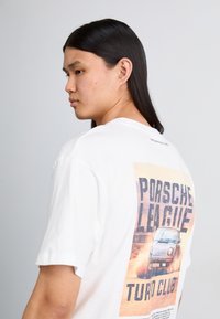 Hvid bomulds t-shirt med et stort grafisk motiv på ryggen, der viser teksten "Porsche League Turbo Club" og en illustration af en vintage bil.