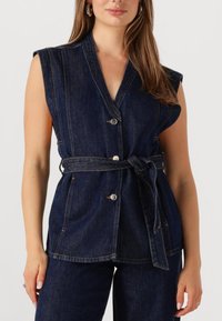 Donkerblauwe spijkerharness zonder mouwen met een wrapdesign, voorzien van twee knopen aan de voorkant, een strikdetail in de taille en zichtbare stiksels.