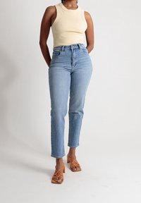 Kermankoinen hihaton paita, korkeavyötäröiset vaaleansiniset farkut ja oranssit remmikengät. Farkuissa on suora lahje ja minimaaliset ompeluseikat.