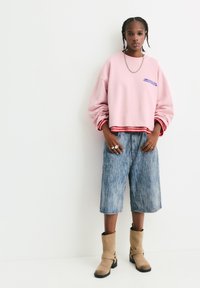 Felpa rosa oversized con testo blu sul petto, orlo a righe rosse; pantaloni shorts di jeans azzurri a fantasia; stivali beige.