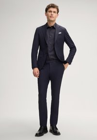 Marineblauer Anzug mit tailliertem Sakko und schmal geschnittenen Hosen, kombiniert mit einem dunklen Hemd mit Knöpfen. Glänzende schwarze Anzugschuhe runden den Look ab.