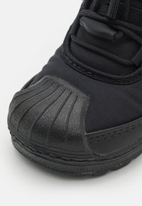 Svart sneaker med texturerat gummitåhätta och ovandel i tyg, med snörning och detaljerad sömnad på sulan och sidan.