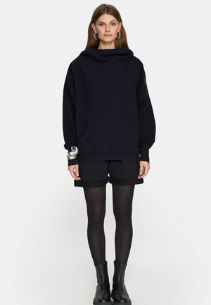 Kobieta stojąca w oversize'owej czarnej bluzie z kapturem, czarnych szortach, przezroczystych czarnych rajstopach, czarnych botkach, srebrnej bransoletce na nadgarstek i kolczykach-kołach.