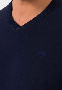 Suéter tejido de color azul marino con diseño de cuello en V. Presenta un sutil logo azul en el lado izquierdo del pecho. Textura suave, bordes del cuello acanalados.