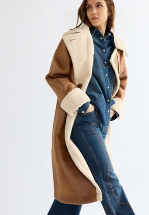 Femme portant un long manteau en peau retournée marron et crème sur une chemise en denim bleu et un jean assorti, les mains dans les poches.