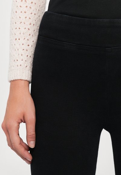 Mustat legginssit, joissa on sileä pinta ja korkea vyötärö, osittain näkyvä käsi kevyessä neulotussa villatakeissa, joissa on avoin kuvio.