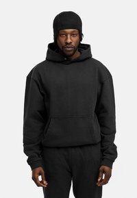 Schwarzer Baumwollhoodie mit einer Fronttasche, verstellbarem Kordelzug und gerippten Bündchen. Getragen mit passenden schwarzen Hosen, vor einem weißen Hintergrund präsentiert.