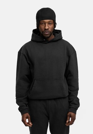 Black cotton hoodie with a front pocket, adjustable drawstring, and ribbed cuffs. Worn with matching black pants, displayed against a white background.