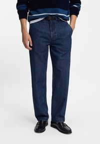 Jean en denim bleu foncé, coupe droite, style cinq poches, tissu de poids moyen, porté avec une ceinture noire et des mocassins noirs.