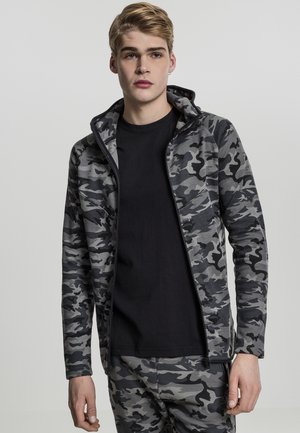 Chaqueta de camuflaje con cremallera en tonos grises, hecha de tela suave, cuenta con capucha y base en negro contrastante. El modelo lleva pantalones a juego.