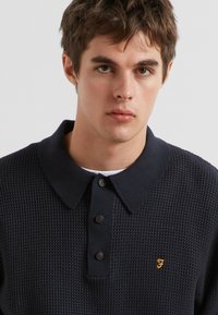 Navy-textured polo shirt met een kraag, drie-knopssluiting en een klein oranje logo op de borst. Gemaakt van ademend materiaal.