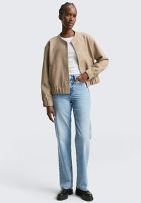 Kvinna klädd i en beige knäppt jacka, vit skjorta, ljusblå jeans och svarta stövlar, står med ena handen i fickan mot en enfärgad bakgrund.