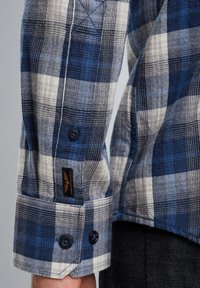 Blauwe en witte geruite flanellen overhemd met lange mouwen, voorzien van een knoopsluiting bij de manchetten en een stoffen label op de mouw. Gelaagde textuur.