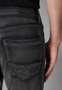 Crne traperice od denima s teksturiranim završetkom, s dizajnom stražnjih džepova i oznakom "DENIM SUPPLY Co." na pojasu.