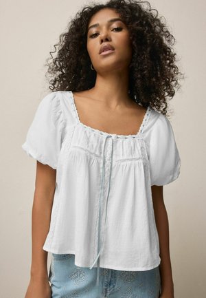 Frau mit lockigem Haar, die eine locker sitzende weiße Bluse mit Puffärmeln und quadratischem Ausschnitt trägt, kombiniert mit hellblauen gemusterten Jeans.