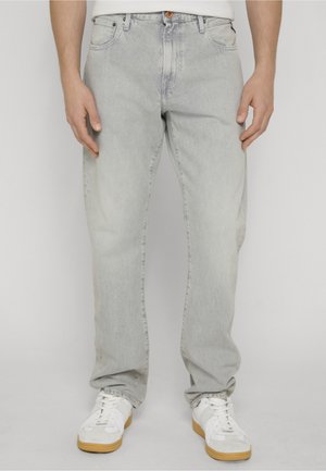 Hombre con vaqueros gris claro y zapatillas blancas de pie contra un fondo claro y liso, con las manos relajadas a los lados.