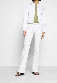 Nainen, jolla on yllään valkoinen farkkutakki ja housut, oliivinvihreä t-paita ja mustat avonaiset korkokengät, seisoo yksinkertaista harmaata taustaa vasten.