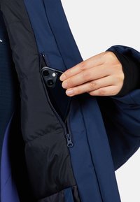 Giacca imbottita blu navy con tasca con cerniera. Una mano sta inserendo uno smartphone nero nella tasca, evidenziando il design funzionale della giacca.