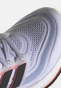 Biały but sportowy z cholewką z siateczki o teksturowanych wzorach, szarymi akcentami i czarnymi detalami na półprzezroczystej podeszwie. Sznurówki są białe.