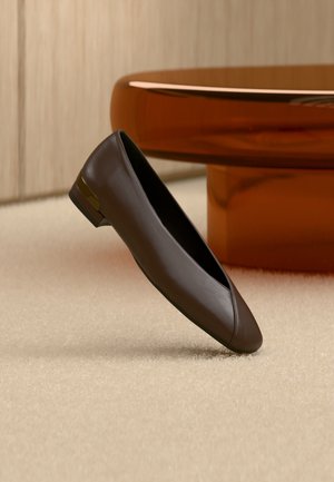 Chaussure plate en cuir brun foncé avec un petit talon et un accent doré, posée sur un tapis beige près d’une table ronde de couleur ambre.