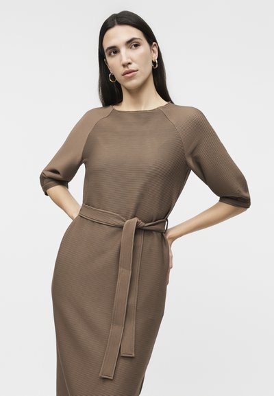 Γυναίκα με μακριά σκούρα μαλλιά που φορά χρυσούς κρίκους στα αυτιά και ένα καφέ εφαρμοστό ριμπ φόρεμα με ζώνη στη μέση, ποζάροντας με τα χέρια στη μέση.