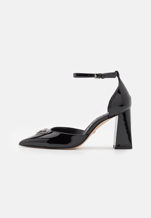 Zapato de tacón con punta alargada en charol negro, con correa en el tobillo y hebilla plateada, con tacón grueso y detalle decorativo en la punta.