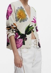 Floral-patterned cardigan in cream featuring multicolored flowers, ribbed cuffs, and four front buttons. Paired with white distressed jeans.