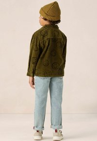 Groene corduroy jas met een glimlachend gezicht print, lichtblauwe jeans, witte sneakers met rode accenten en een beige gebreide beanie.