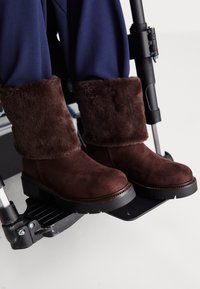 Sötétkék nadrágot viselő lábak és barna szőrmével bélelt csizmák pihennek egy tolószék lábtartóján, fehér háttér előtt.