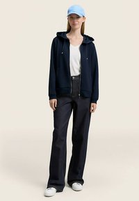 Tamsiai mėlynas džemperis su užtrauktuku ir raišteliais, balta marškinėliai, tamsios mėlynos plačios džinsinės kelnės ir šviesiai mėlyna kepurė. Modelis avėdams baltus sportinius batelius.