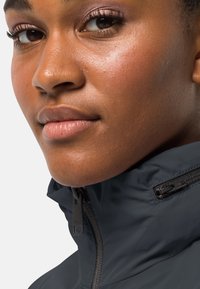 Primo piano di una giacca grigio scuro con colletto alto, caratterizzata da una superficie texturizzata e dettagli in cerniera ben visibili. È visibile un tono di pelle uniforme.