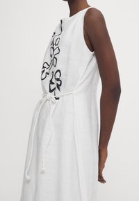 Valkoinen pellavapuku, jossa on mustaa kukkamusteita edessä, ilman hihoja ja nyöri vyötäröllä valkoisella köydellä.