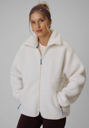 Femme aux cheveux blonds et aux yeux bleus portant une veste polaire blanche zippée et un bandeau marron, les mains dans les poches de la veste sur fond gris.