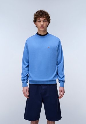 Young man with curly hair wearing a light blue long-sleeve sweater and dark blue knee-length shorts, standing against a plain light background.