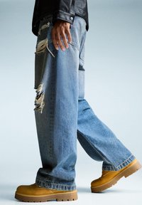 Person wearing ripped wide-leg blue jeans, tan work boots, and a black leather jacket, shown from mid-thigh down against light background.