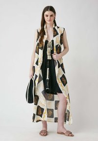 Flowing, sleeveless kimono in cream with black and gold abstract patterns. Paired with a black dress, tassel details, and neutral sandals.