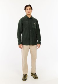 Muškarac stoji u tamnozelenoj flis jakni, bež hlačama i zelenim planinarskim cipelama na bijeloj podlozi.