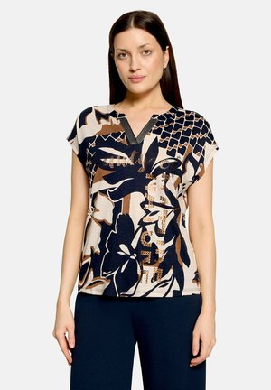 Kvinde iført en kortærmet beige, sort og brun top med blomsterprint og V-hals samt mørkeblå bukser mod en ensfarvet baggrund.