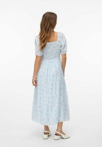 Robe florale bleu clair avec des manches bouffantes et un dos smocké. Présente une jupe mi-longue et de petits motifs de fleurs roses. Portée avec des talons beiges.