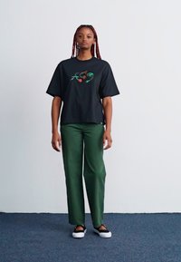 Juodas per didelis T-shirt'as su ryškiu gėlių siuvinėjimu, derinamas su plačiomis žaliomis kelnių ir juodai baltais bateliais be raištelių.