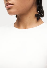 Κοντινό πλάνο της κάτω πλευράς του προσώπου ενός ατόμου, του λαιμού και της περιοχής των ώμων με πλεγμένα μαλλιά, που φοράει μια απλή λευκή μπλούζα με στρογγυλή λαιμόκοψη.