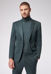 Ciemnozielony garnitur dwuczęściowy z teksturowanej tkaniny, dopasowana marynarka i golf pod spodem. Posiada dwie przednie kieszenie oraz subtelne detale akcentujące.