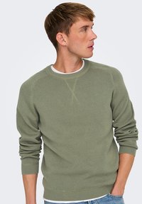 Maglione verde oliva con scollatura a girocollo, polsini e orlo a costine, e un motivo a maglia testurizzato. Indossato sopra una camicia bianca. Vestibilità casual.
