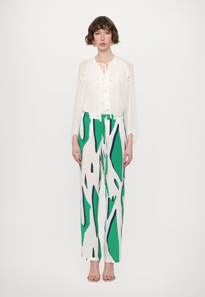 Blouse blanche à volants avec un lien au cou, associée à un pantalon large à motifs verts et blancs. Talons légers.