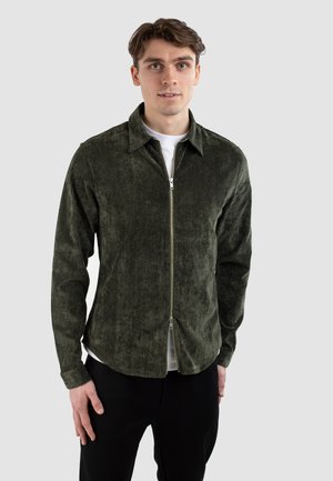 Dunkelgrüne, strukturierte Jacke mit Reißverschluss, spitzem Kragen und langen Ärmeln, kombiniert mit einem weißen Hemd darunter.